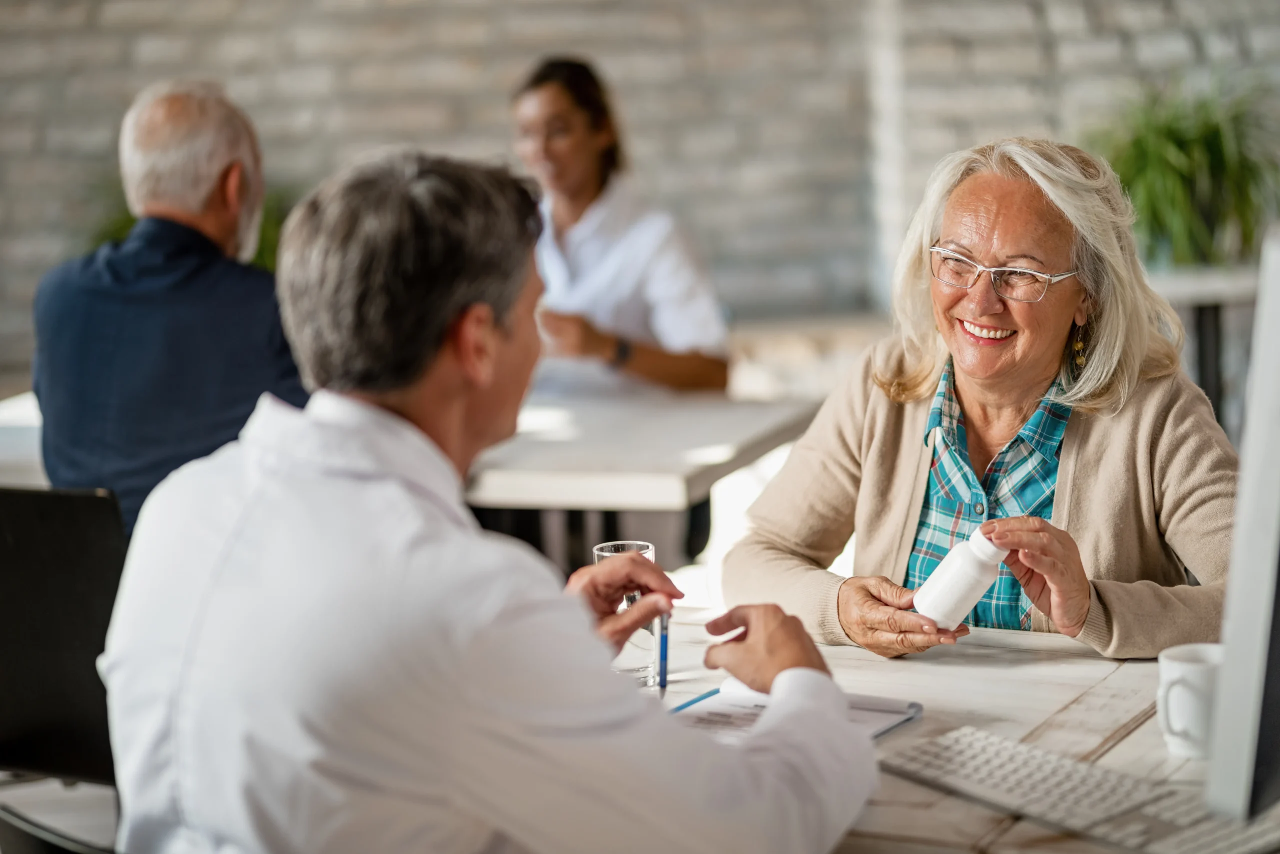 An older woman holds prescriptions and discusses with a doctor that sits at keyboard. Another doctor-patient pair sits in the background.