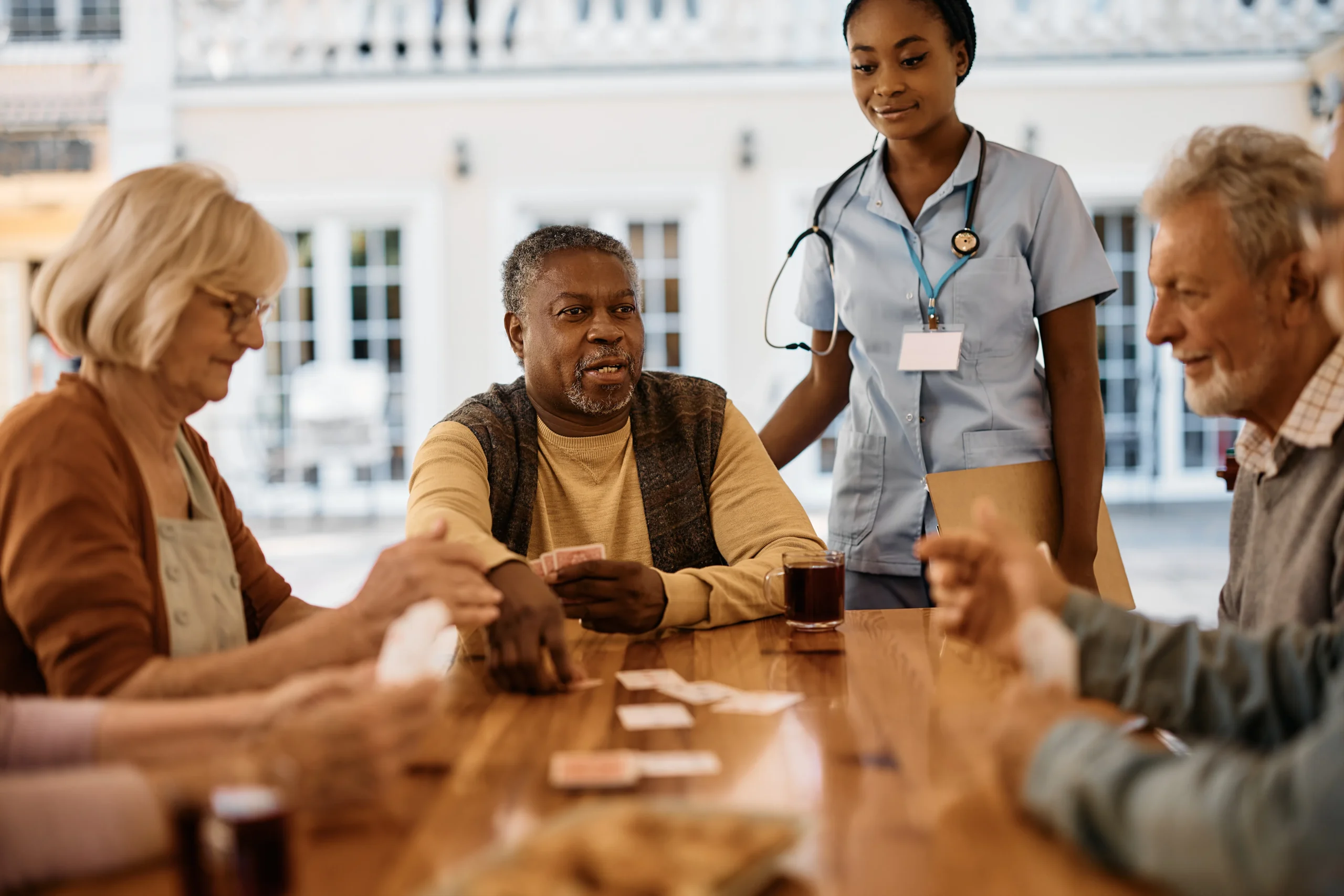 A multiracial group of senior friends playing cards at an assisted care facility with nurse watching and smiling.